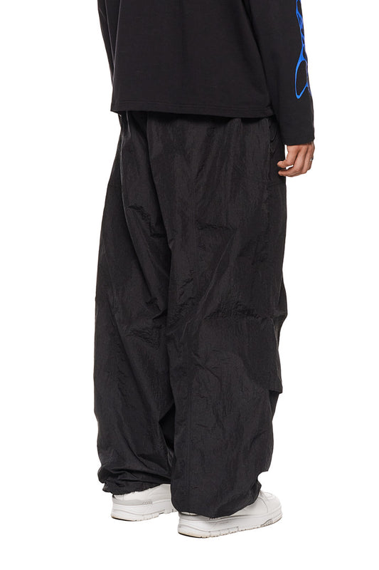 Eine Person trägt OLAKALA Parachute Pants Black mit elastischem Kordelzug, Eingriffstaschen und gesticktem Synergy-Logo, ideal für einen lässigen, bequemen Look.