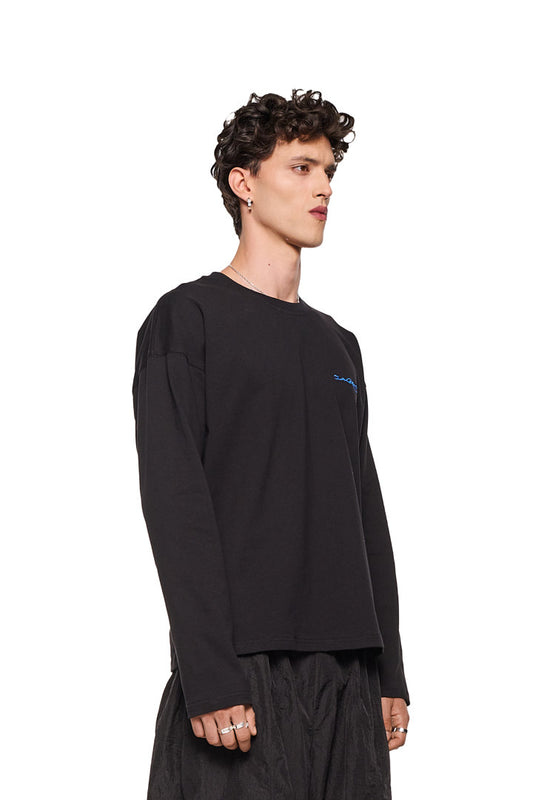 Mann trägt Synergy Embroidered Longsleeve Black mit gesticktem Logo auf der Brust, oversized Passform; Detailaufnahme einer Hand mit Ring.
