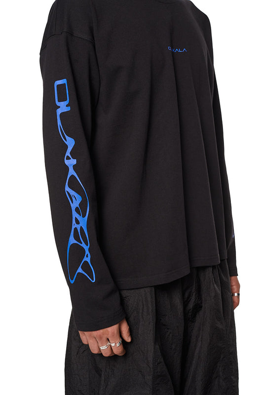 Person trägt das Synergy Longsleeve Shirt Black mit OLAKALA Logo auf der Brust und Synergy-Logos auf den Ärmeln, in oversized Passform.