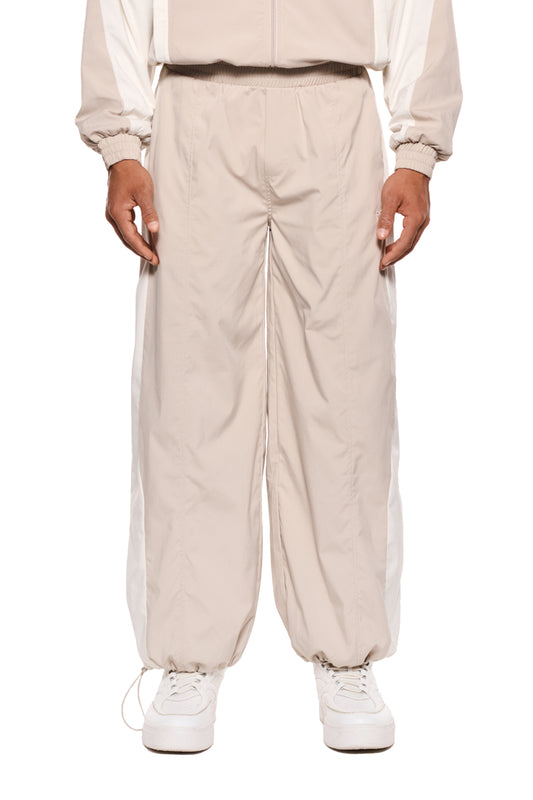 Person trägt Sports Tracksuit Pants Sand mit Teilungsnähten und Reißverschlusstaschen, elastischem Kordelzug, gesticktem OLAKALA Sports Icon, lockere Passform, robustes Rip-Stop-Gewebe.