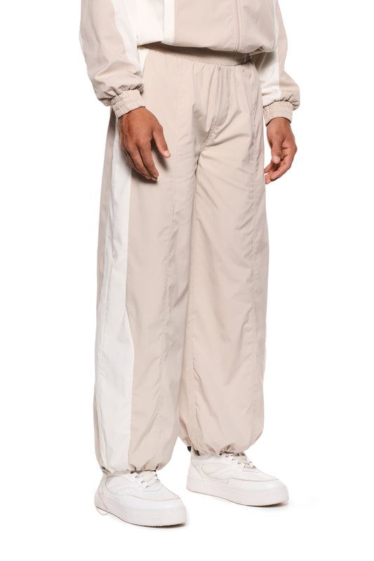 Person trägt OLAKALA Sports Tracksuit Pants Sand mit Teilungsnähten, elastischem Kordelzug und Reißverschlusstaschen, ergänzt durch gesticktes Logo und robustes Rip-Stop Gewebe.
