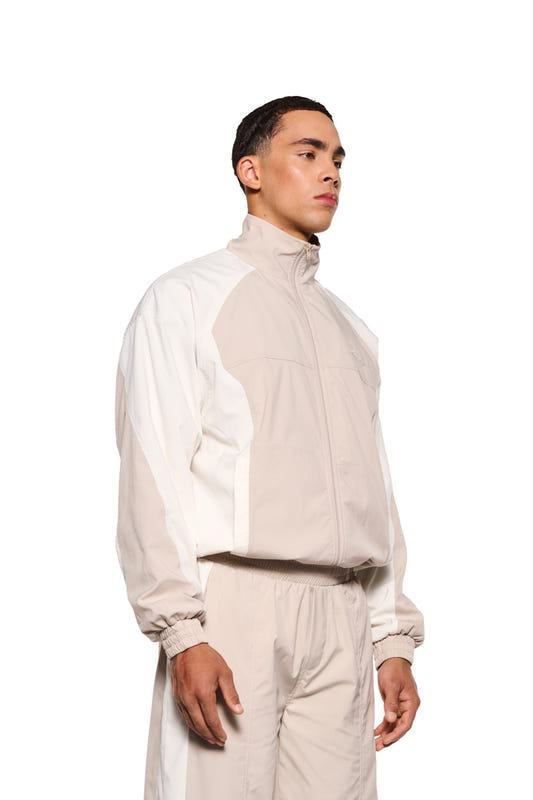 Mann trägt OLAKALA Sports Tracksuit Jacket Sand mit elastischem Bund, Reißverschlusstaschen und gesticktem Logo auf der Brust. Robustes Rip-Stop-Gewebe, lockere Passform.