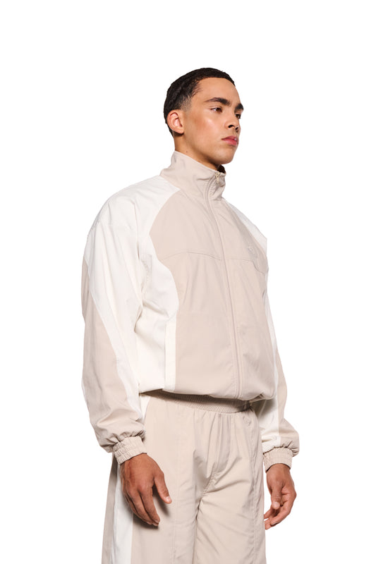 Mann trägt OLAKALA Sports Tracksuit Jacket Sand mit elastischem Bund, Reißverschlusstaschen und gesticktem Logo auf der Brust. Robustes Rip-Stop-Gewebe, lockere Passform.