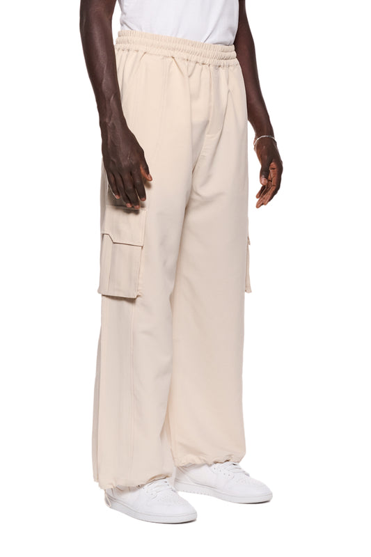 Person trägt Synergy Cargo Pants Sand mit elastischem Kordelzug, Eingriffstaschen und Cargo-Taschen mit Magnetverschluss. Sichtbare Teilungsnähte und gesticktes Logo.