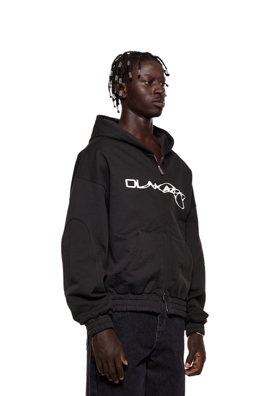 Mann trägt Synergy Zip Hoodie Black mit 2-Wege-Reißverschluss und Synergy-Logo auf der Brust. Oversized Unisex-Design, Teilungsnähte an den Ärmeln sichtbar.