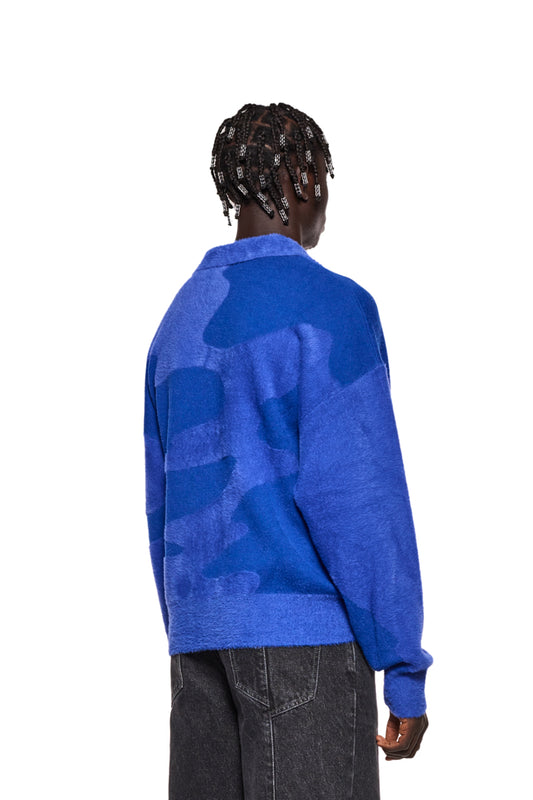 Person trägt Synergy Glitched Fuzzy Polo Blue mit Polo-Kragen, gesticktem Synergy-Logo auf der Brust und Ripp-Bündchen, unisex Design.