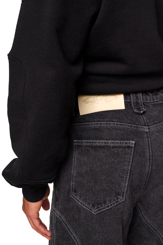 Person trägt Alien Intel Black Denim Pants mit 5-Pocket-Style, geprägtem OLA KALA PU-Patch und geradem Bein für lockere Passform.