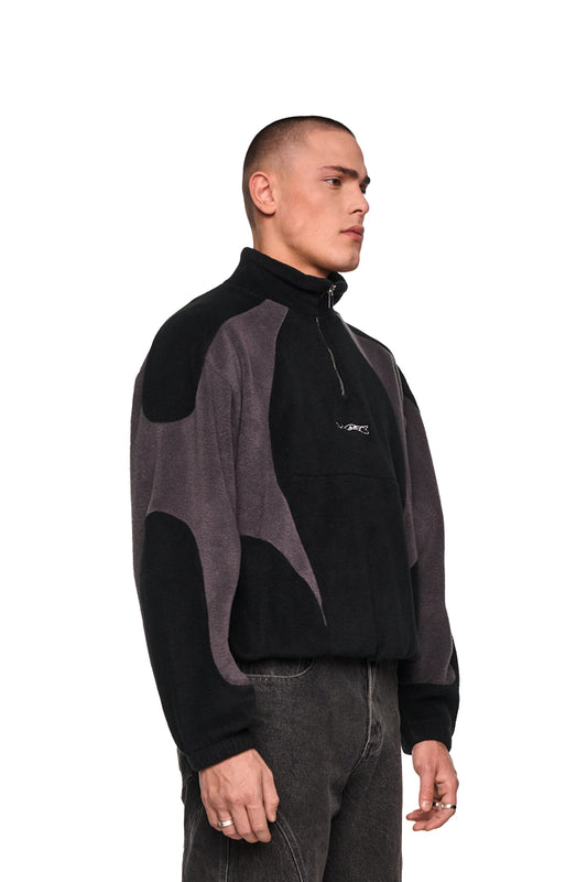 Mann im Profil trägt Alien Intel Fleece Halfzip Black mit hohem Kragen, Brustreißverschluss und gesticktem „Synergy“-Logo, zeigt elastische Bündchen und Oversized-Passform.