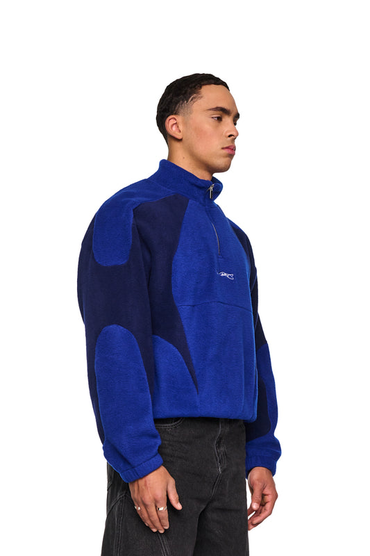 Mann trägt Alien Intel Fleece Halfzip Blue mit hohem Kragen, Brustreißverschluss und gesticktem Synergy-Logo, gezeigt im Profil.