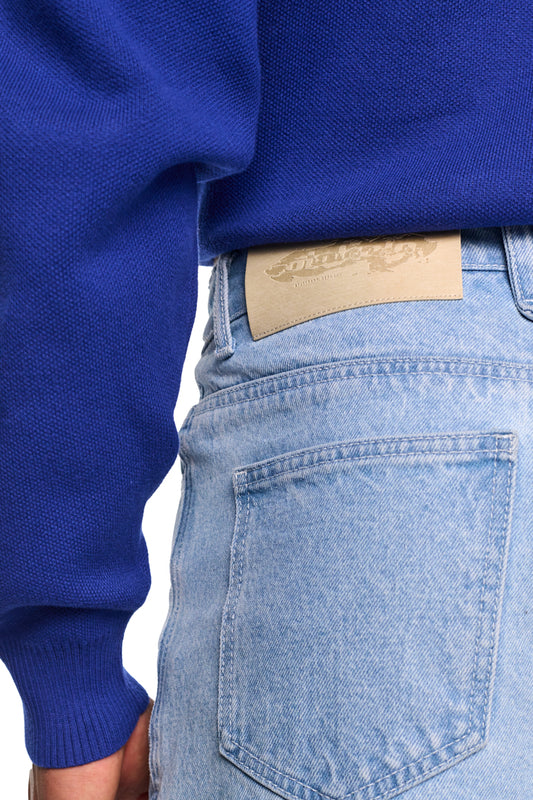 Person trägt Alien Intel Blue Denim Pants mit geradem Bein und lockerem Schnitt, betont durch Teilungsnähte und 5-Pocket-Style. Geprägtes OLA KALA PU-Patch sichtbar.