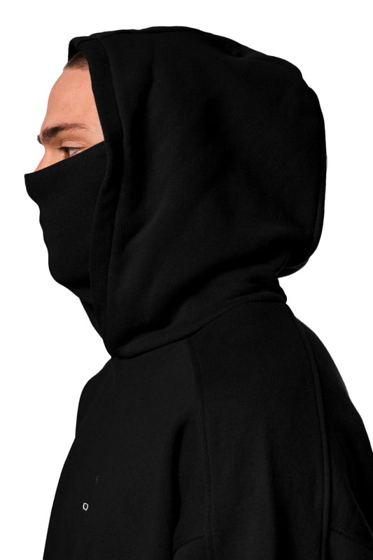 Person in schwarzem Discreet Mask Hoodie Black von OLAKALA mit Ripp-Bündchen, elastischem Saum und gesticktem Defect Identity Icon auf der Brust.