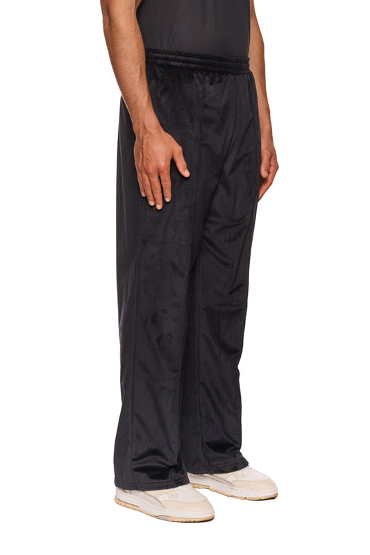 Person trägt Sports Velour Jogger Pants Black mit offener Beinabschluss und Eingriffstaschen, zeigt gesticktes OLAKALA Sports Icon auf linkem Bein.