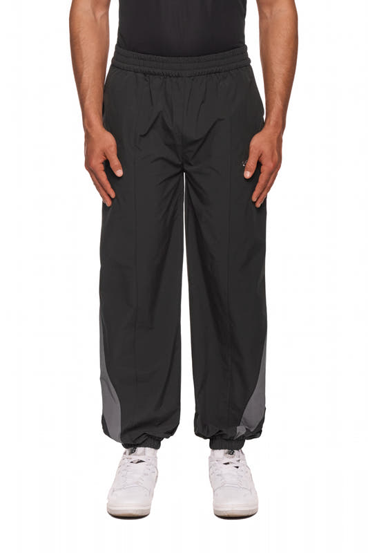 Person trägt OLAKALA Sports Trackpants Black mit elastischem Bund, kontrastfarbene Einsätze und gesticktem Logo auf linkem Bein, kombiniert mit weißen Sneakers.