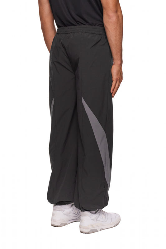 Person trägt schwarze Sports Trackpants mit gesticktem OLAKALA Sports Logo, elastischen Bündchen und kontrastierenden Stoffeinsätzen, ergänzt durch weiße Sneakers.