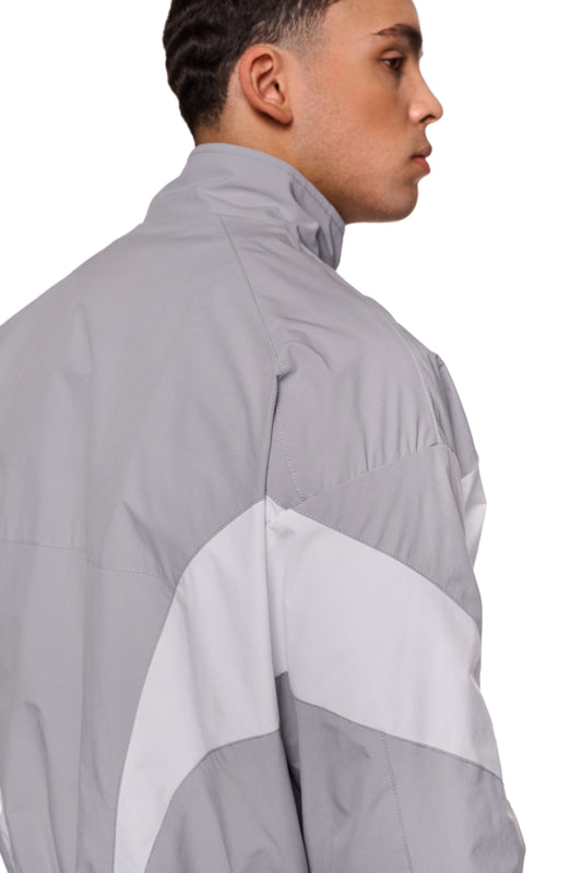 Mann in grauer Sports-Trackjacke, robustes Rip-Stop-Gewebe, atmungsaktives Mesh-Futter, sportliche Stoffeinsätze, elastische Bündchen, OLAKALA Sports Logo auf Brust sichtbar.
