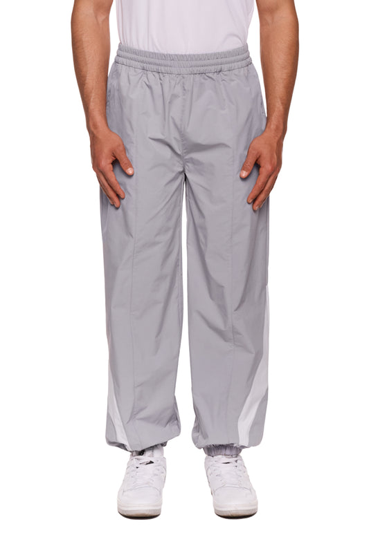 Person trägt graue Sports Trackpants von OLAKALA mit Rip-Stop Gewebe, kontrastierenden Stoffeinsätzen und gesticktem Logo auf dem linken Bein. Hände in den Taschen.