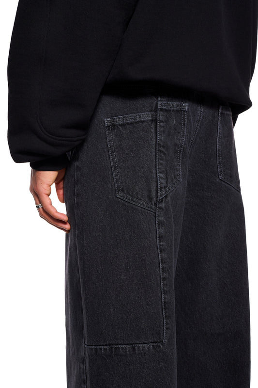 Person trägt CC Black Denim Pants Baggy mit versetztem Hosenbund und Mittelfinger-Patch, zeigt eine Gesäßtasche und einen OLAKALA-Jeansknopf.