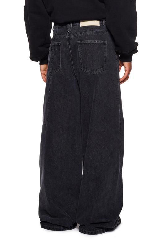 Person trägt CC Black Denim Pants Baggy mit „Mittelfinger“ Patch und OLAKALA Logo, lockere Passform, betont durch versetzte Nähte und Tasche.