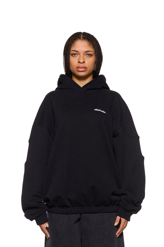 Woman wears CC Hoodie Black with embroidered OLAKALA logo, dividing seams on sleeves, ribbed cuffs and elastic hem. Oversized unisex fit.