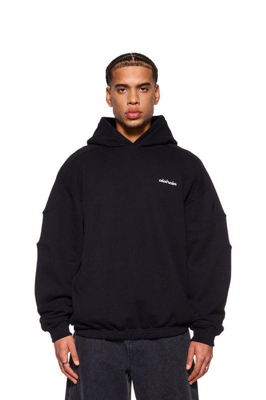 Mann trägt CC Hoodie Black mit gesticktem OLAKALA Logo, Oversized-Passform, Teilungsnähten und Ripp-Bündchen an den Ärmeln. Unisex-Design.