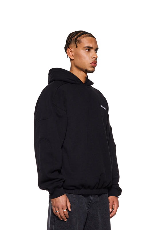 Mann trägt CC Hoodie Black mit gesticktem OLAKALA Logo auf der Brust und Teilungsnähten an den Ärmeln. Oversized Passform, unisex, mit Ripp-Bündchen.