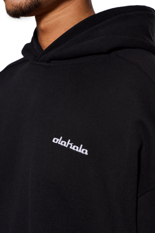 Person wears the CC Hoodie Black with embroidered OLAKALA logo, dividing seams on the sleeves and ribbed cuffs, in an oversized unisex fit.