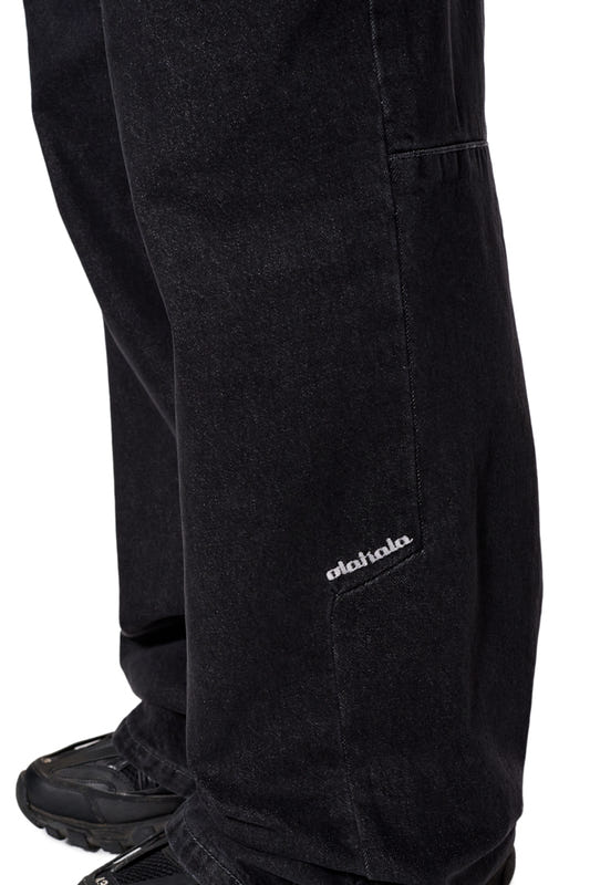 Nahaufnahme der CC Black Loose Fit Denim Pants mit versetzter Seitennaht und gesticktem OLAKALA-Logo auf der linken Vorderhose.