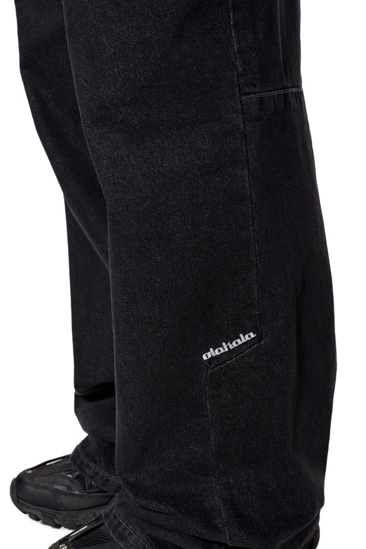 Nahaufnahme der CC Black Loose Fit Denim Pants mit versetzter Seitennaht und gesticktem OLAKALA-Logo auf der linken Vorderhose.