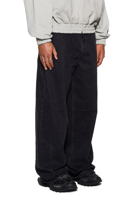 Person trägt CC Black Loose Fit Denim Pants mit versetzter Seitennaht und OLAKALA Details, inklusive „Mittelfinger“ Patch und geprägtem Jeansknopf.