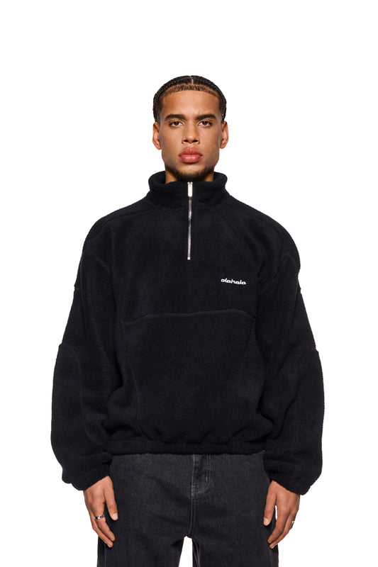 Mann trägt CC Fleece Halfzip Black mit gesticktem OLAKALA Logo, elastischem Bund, hohem Kragen und Reißverschluss, Oversized Fit, sichtbar sind Jacke und Jeans.