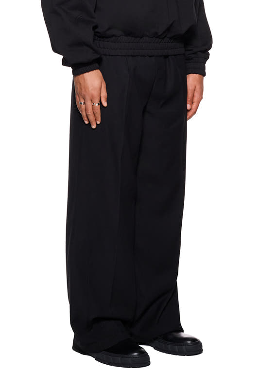 Person wears Gills dress pants with French pockets, pleats and embroidered OLAKALA logo. Visible details: black pants, hand with silver rings, black shoes.