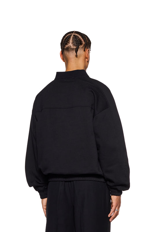Person trägt CC Polo Longsleeve Black mit Polo-Kragen, Knopfleiste, elastischen Bündchen und gesticktem OLAKALA-Logo auf der Brust. Oversized Passform, unisex.