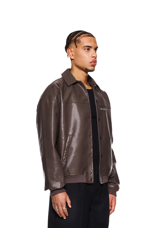 Mann in brauner CC Leather Jacket mit Reißverschluss, Leistentaschen, gesticktem OLAKALA Logo und veganem Lederimitat. Oversized, unisex Design.