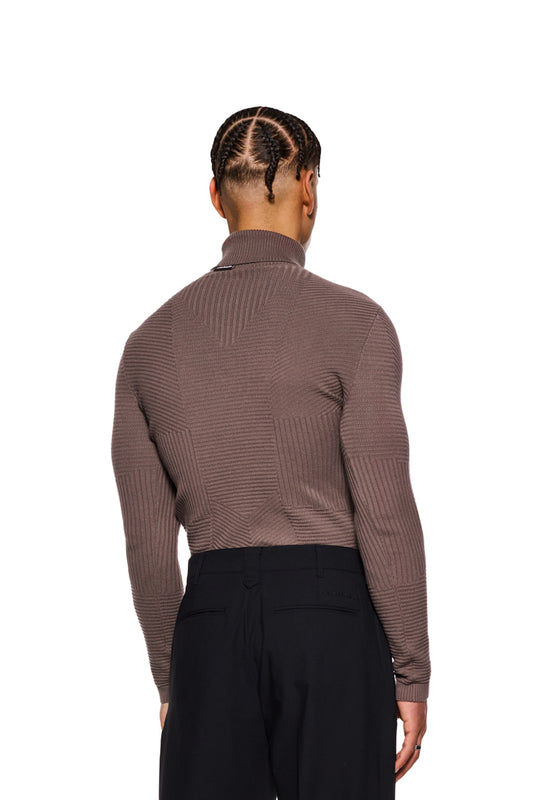 Mann in einem eng anliegenden, braunen Anatomy Turtleneck mit Rollkragen und OLAKALA-Tag am Kragen, trägt schwarze Hosen.