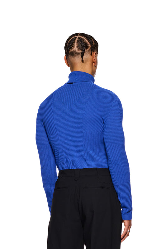 Mann trägt Anatomy Turtleneck Blue, einen eng anliegenden Strickpullover mit Rollkragen und OLAKALA-Tag am Kragen, kombiniert mit schwarzen Hosen.