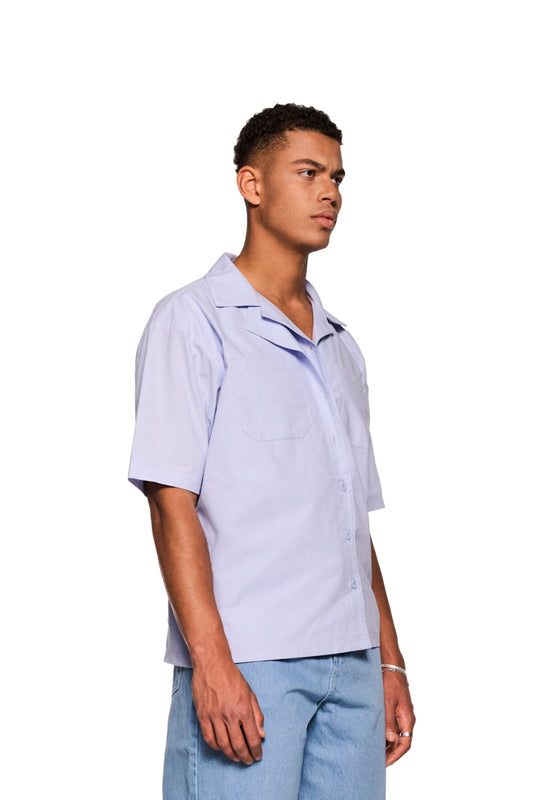 Mann im Layered Short Sleeve Shirt Blue mit kubanischem Kragen und Brusttaschen, gesticktes OLAKALA-Logo sichtbar, lockere Passform, unisex.