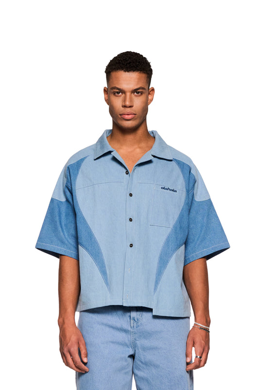 Mann trägt Echoes Denim Shirt mit kubanischem Kragen und Brusttasche. Lockerer Schnitt, markante Teilungsnähte, gesticktes OLAKALA-Logo.