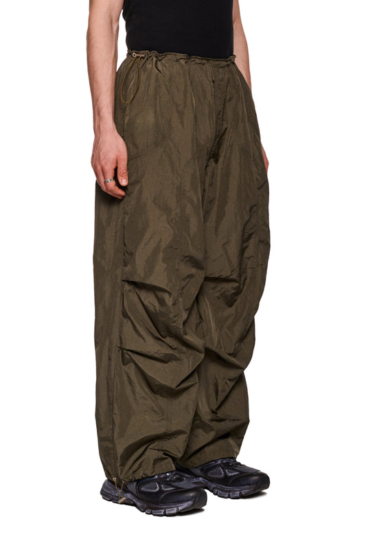 Person trägt Parachute Pants Khaki mit elastischem Kordelzug und gesticktem Synergy-Logo, betont durch Seitentaschen und Paspeltasche. Lockere Passform, strapazierfähiges Material.