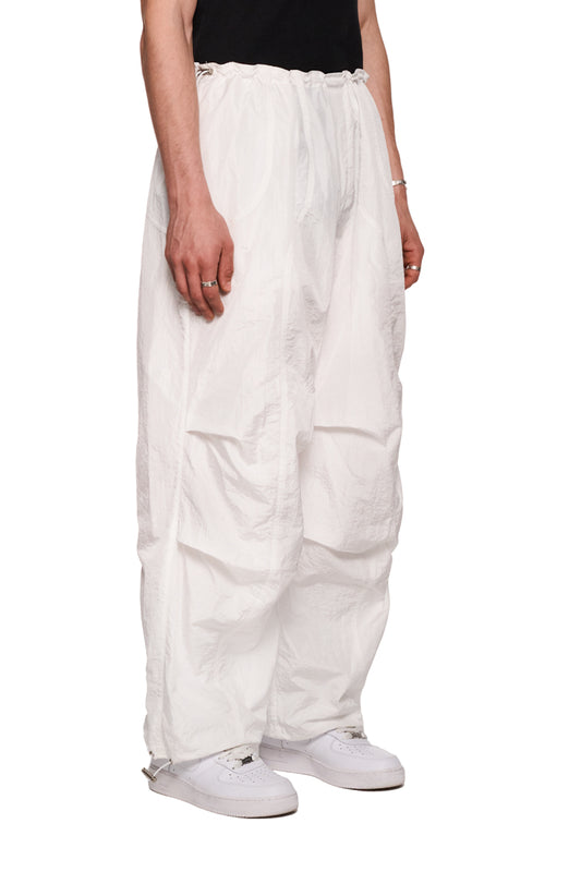 Person trägt weiße Parachute-Pants mit OLAKALA-Kordelzug, Eingriffstaschen und gesticktem Synergy-Logo, betont durch doppelt abgesteppte Teilungsnähte.