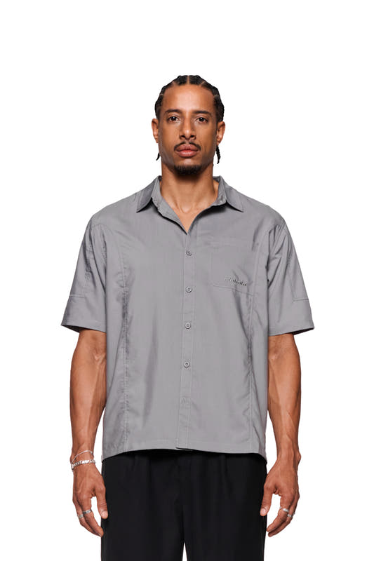 Man wears short-sleeved CC shirt in gray with OLAKALA logo on the breast pocket, front button placket and loose fit.