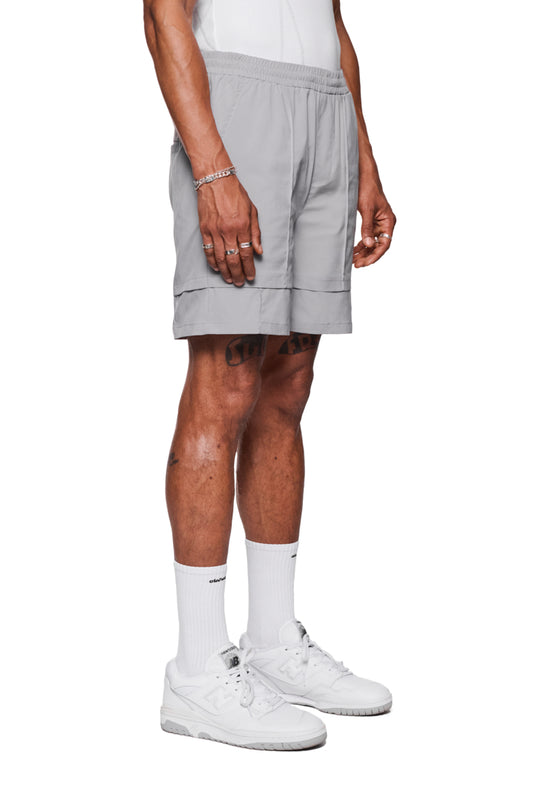 Mann trägt Track Shorts Gray mit elastischem Bund und gesticktem OLAKALA-Logo, kombiniert mit weißen Sneakers und Socken. Ideal für Alltag oder als Badehose.