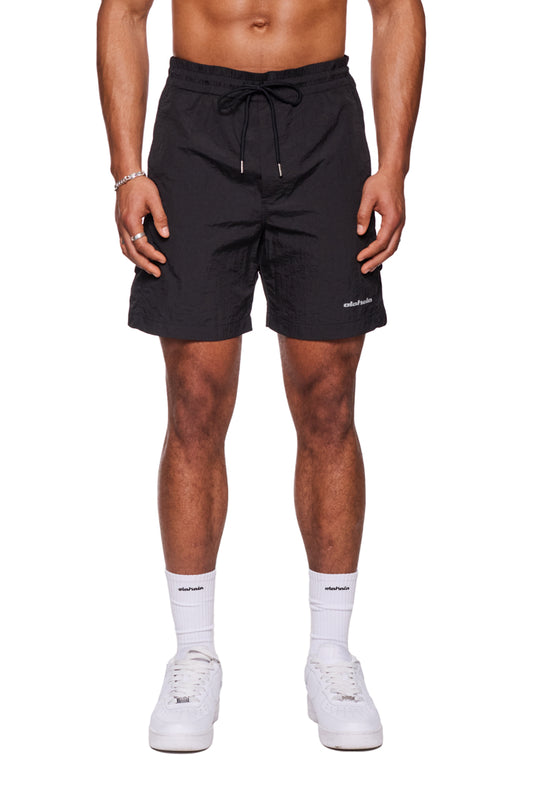 Mann trägt Swim Shorts Black mit Monogramm-Effekt, elastischem Bund und reflektierendem Logo-Print, kombiniert mit weißen Sneakern und sichtbaren Eingriffstaschen seitlich.