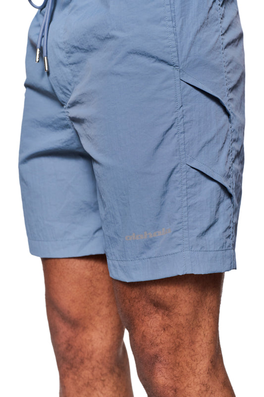 Person trägt Swim Shorts Faded Blue mit Monogramm-Effekt und reflektierendem Logo-Print, elastischem Bund mit Kordelzug, und seitlichen Eingriffstaschen, ideal für den offiziellen OLAKALA® Onlineshop.