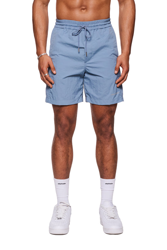 Mann trägt Swim Shorts Faded Blue mit Monogramm-Effekt, elastischer Bund, Kordelzug, reflektierender Logo-Print und seitliche Taschen, kombiniert mit weißen Sneakers.