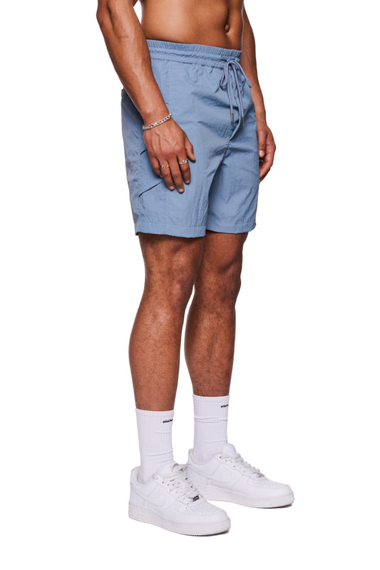 Mann trägt Swim Shorts Faded Blue mit elastischem Bund und Kordelzug, seitlichen Eingriffstaschen und reflektierendem Logo-Print, kombiniert mit weißen Socken und Sneakers.