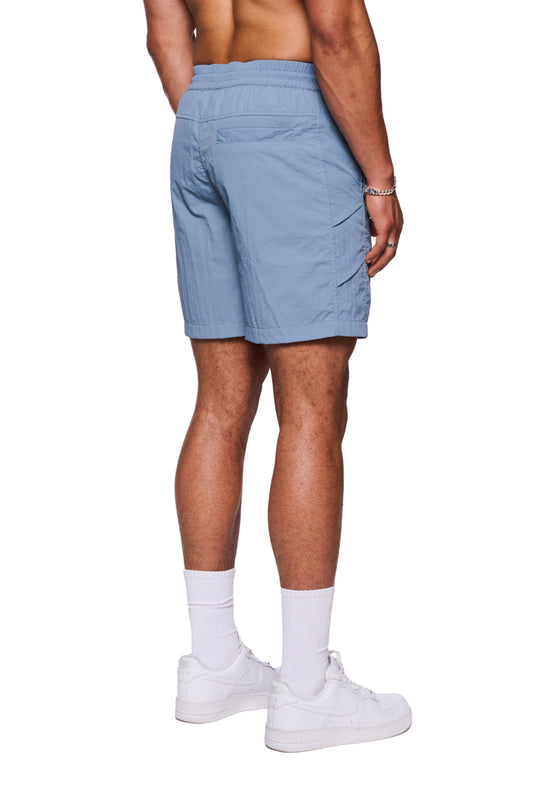 Person trägt Swim Shorts Faded Blue mit elastischem Bund und Kordelzug, seitlichen Eingriffstaschen und reflektierendem Logo-Print, kombiniert mit weißen Sneakers und Socken.