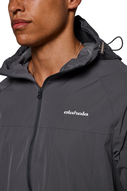 Person trägt Windbreaker Dark Gray mit Kapuze, elastischen Bündchen und gesticktem OLAKALA Logo auf der Brust; wasser- und winddicht, Reißverschlusstaschen vorne.