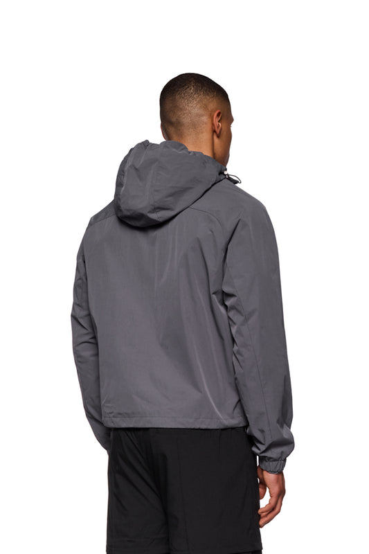 Mann trägt dunkelgrauen Windbreaker mit Kapuze und elastischen Details, sichtbar sind Reißverschlusstaschen und gesticktes OLAKALA-Logo auf der Brust.