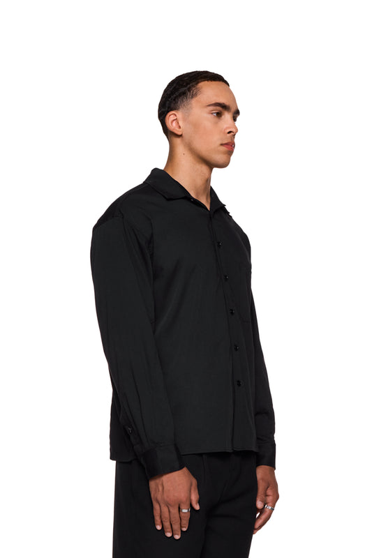 Mann im CC Anatomy Shirt Black mit Knopfleiste, Doppellagigem Kragen und aufgesetzter Brusttasche mit gesticktem OLAKALA Logo, unisex und locker geschnitten.