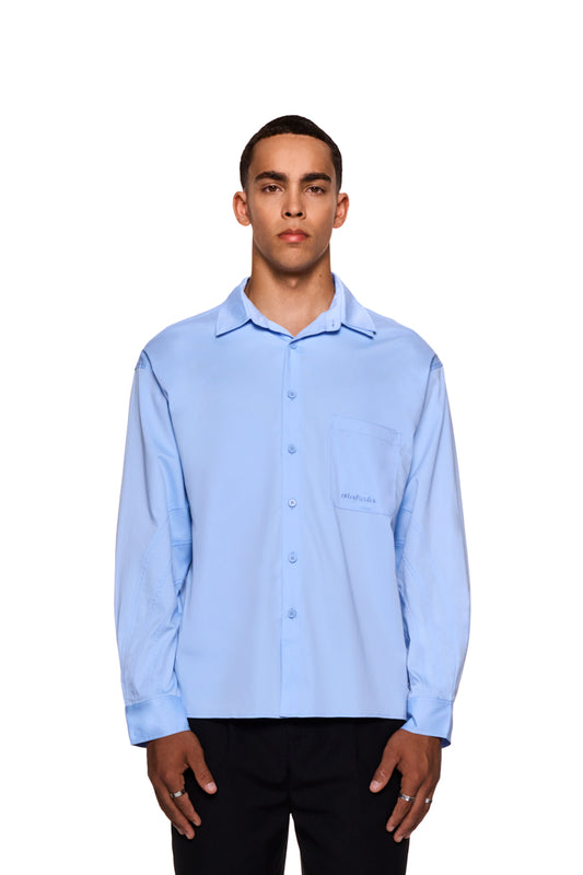Mann trägt das CC Anatomy Shirt Blue mit Knopfleiste, doppellagigem Kragen, Brusttasche mit gesticktem OLAKALA Logo und lockerer Passform.